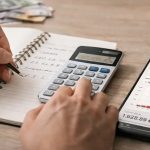 egy férfi számológépen számol jegyzetfüzet mellett, miközben a telefonján pénzügyi online kalkulátor grafikonja látható